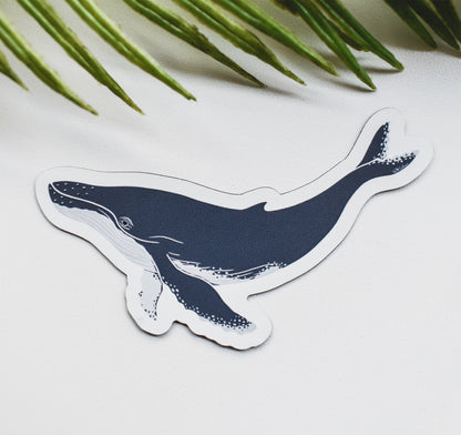 A magnet featuring a stylized humpback whale illustration in dark navy blue and white, placed on a light surface with green palm leaves in the background.