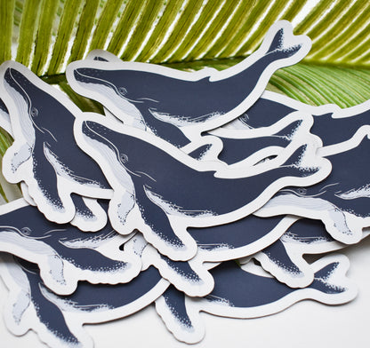 A pile of humpback whale magnets with dark navy blue bodies and white outlines, arranged on a white surface with green palm leaves in the background.