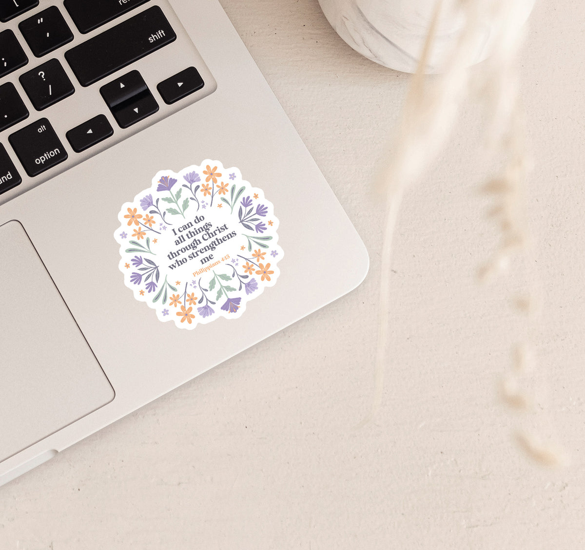 Decorative Christian faith sticker with the Bible verse "I can do all things through Christ who strengthens me, Philippians 4:13" surrounded by hand-drawn purple and orange flowers, located on a silver laptop