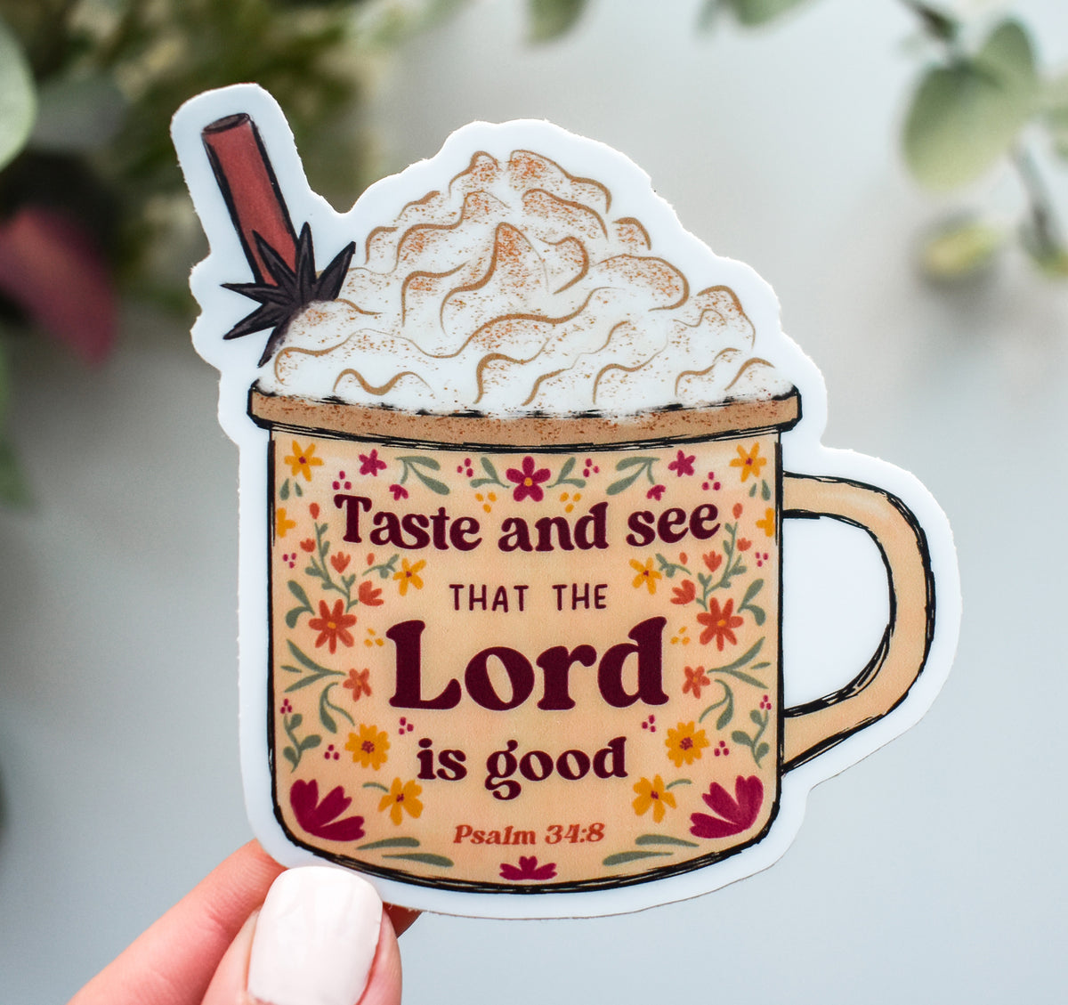 A die-cut sticker shaped like a mug filled with whipped cream and a cinnamon stick, held in a hand. The mug has a floral pattern and the text &